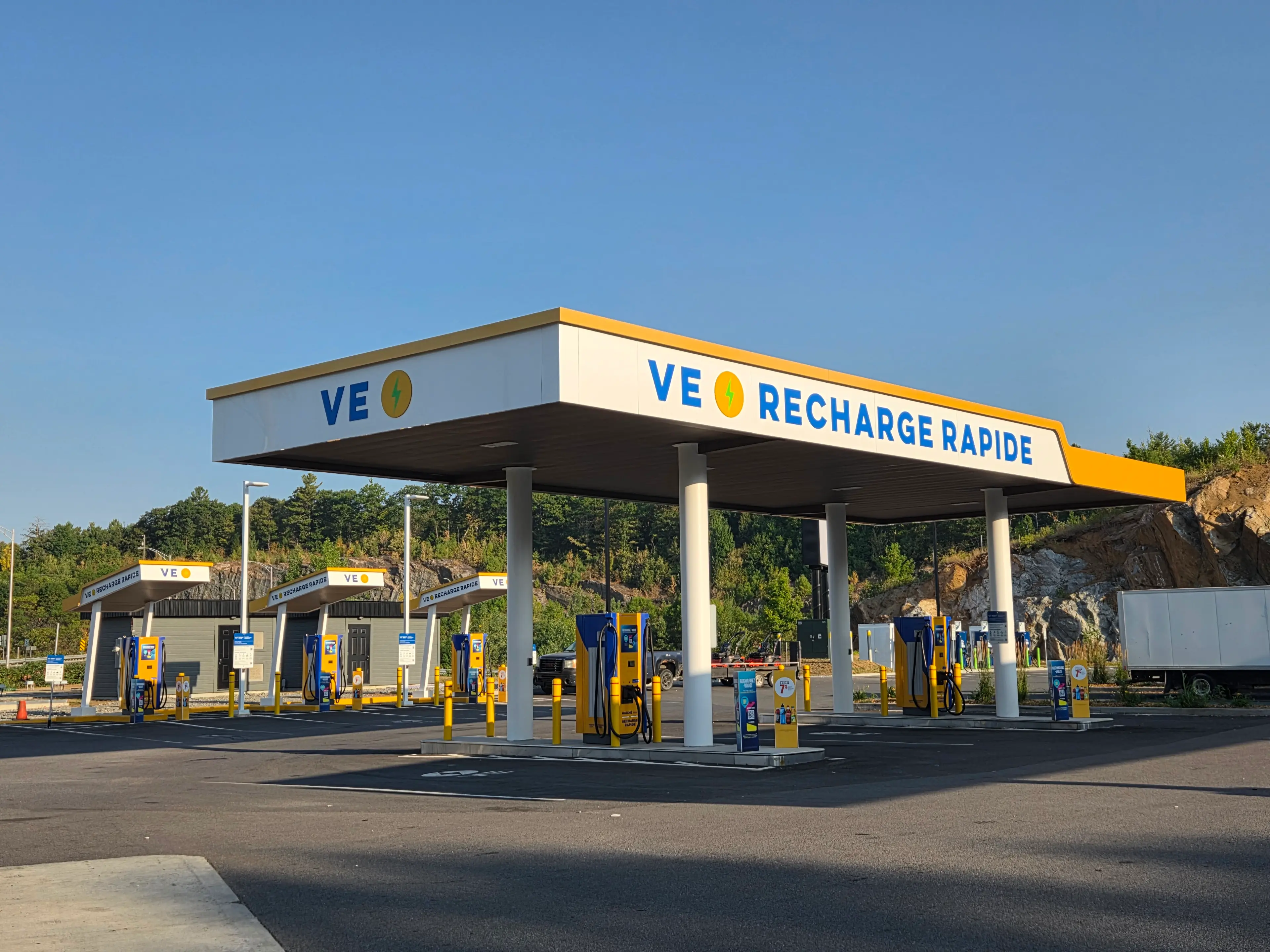 La photo montre une station VE Recharge Rapide avec plusieurs bornes de recharge jaunes et bleues sous un grand auvent blanc. L’enseigne affiche clairement « VE Recharge Rapide » en lettres bleues avec une icône d’éclair jaune. Le site est propre et spacieux, avec plusieurs bornes alignées et un marquage clair au sol.