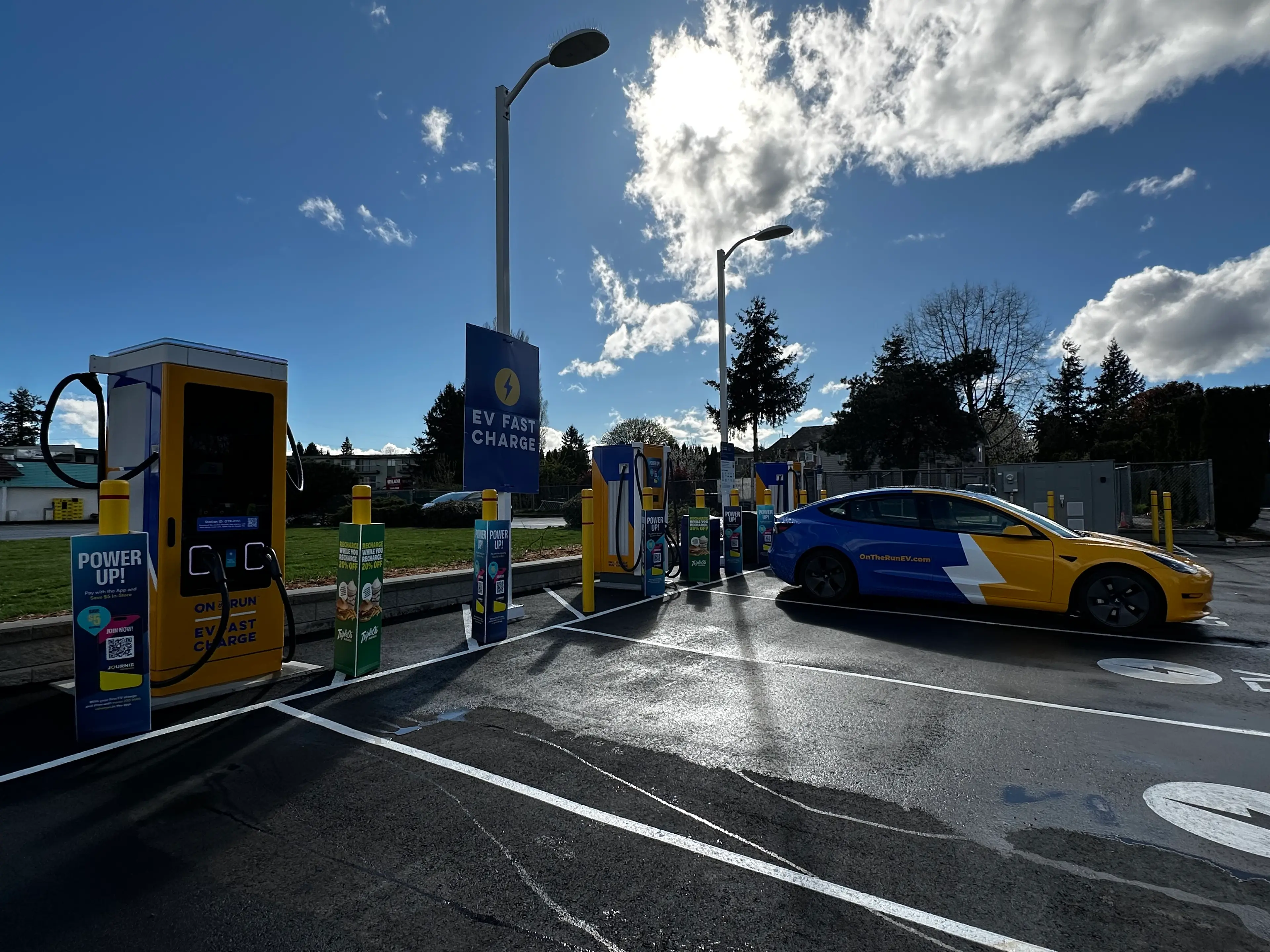 The photo shows an On the Run EV Fast Charge station with yellow and blue chargers. A blue and yellow car with a lightning bolt graphic and OnTheRunEV.com branding is parked and plugged in.