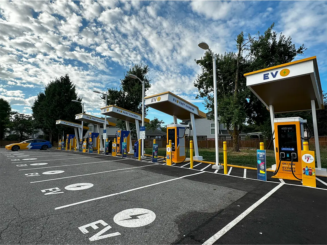 The photo shows an EV Fast Charge station with multiple yellow and blue chargers under white canopies. The parking spots are clearly marked with EV symbols. A blue and yellow EV with a large white lightning bolt graphic is parked at one of the stalls.