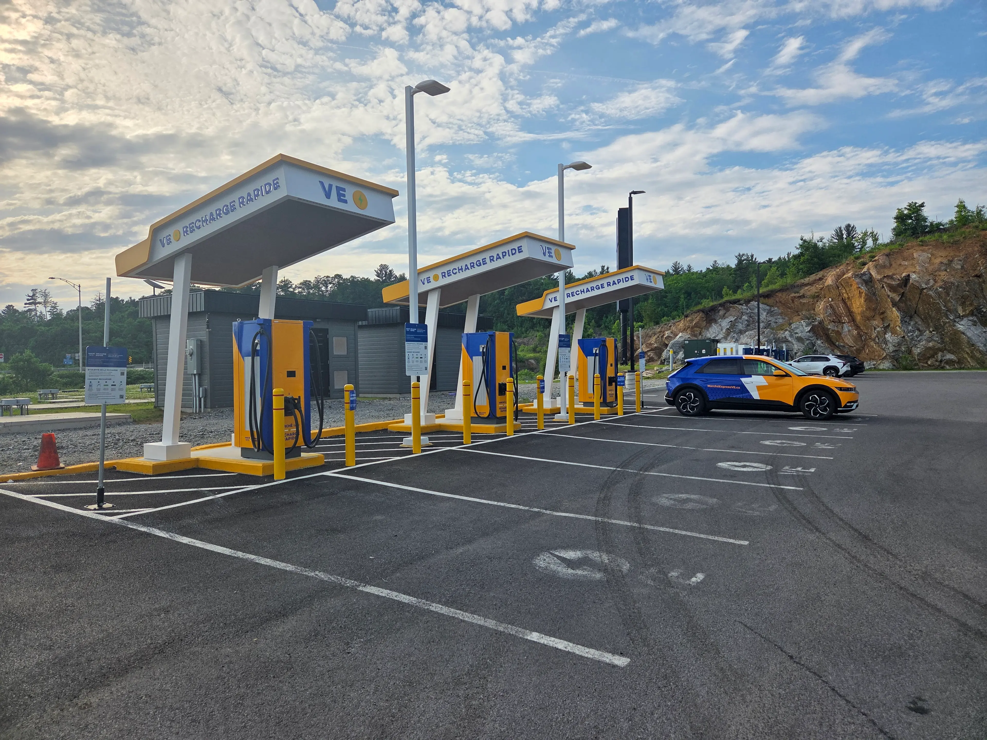 La photo montre une station VE Recharge Rapide avec des bornes de recharge jaunes et bleues. Une voiture électrique bleue et jaune avec un grand éclair blanc et l’adresse MarcheExpressVE.ca est stationnée et branchée.